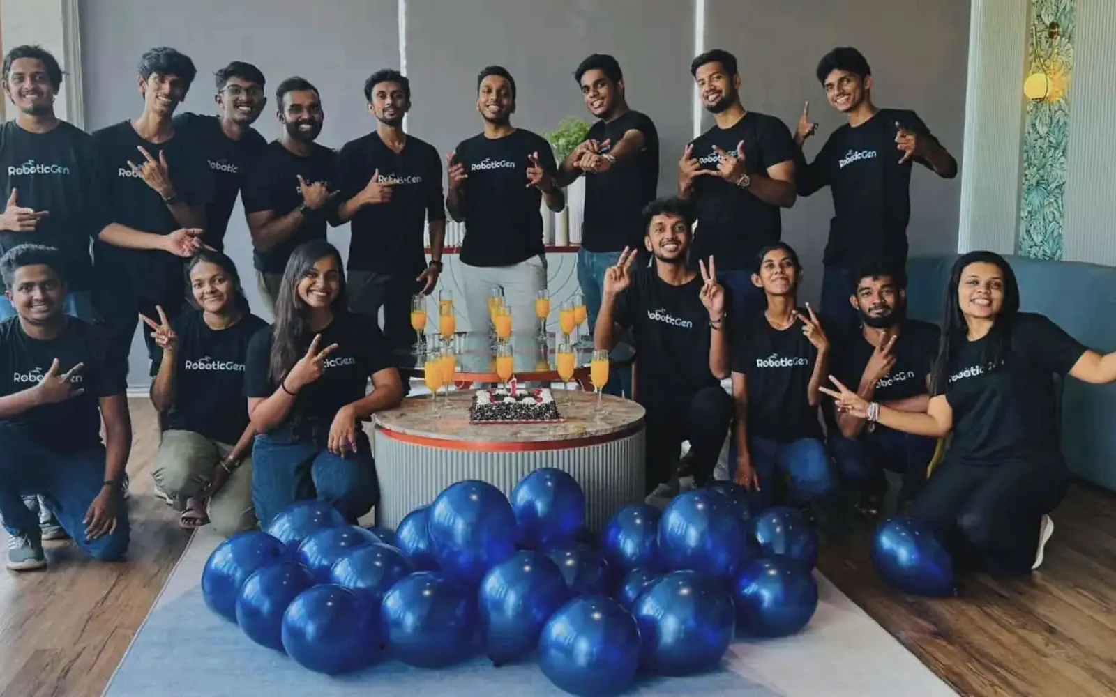 The RoboticGen team in RoboticGen T-shirts, celebrating together around a cake and blue balloons.